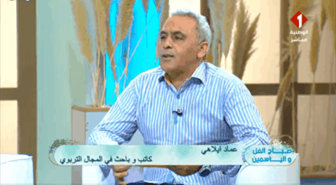 أستاذ عماد إيلاهي في أستوديو القناة الوطنية الأولى، يحمل كتابه حول الإصلاح التربوي، وأمامه ميكروفون يرمز لصوت الكلمة الحرة.